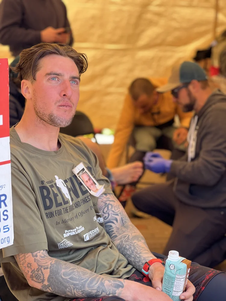 Jared Buchanan at sitting with an energy drink in one hand, at the end of an ultra-thon wearing image of his father on his t-shirt.